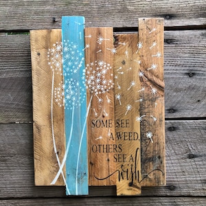 May include: Wooden sign with a blue stripe down the center. The sign features a white painted design of three dandelions with seeds blowing in the wind. The text on the sign reads "Some see a weed, others see a wish."