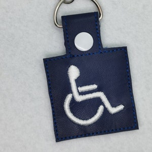 Pode incluir: Um chaveiro de couro azul com um símbolo de acessibilidade em cadeira de rodas bordado em branco. O chaveiro tem um anel de metal prateado e um fecho de metal prateado.