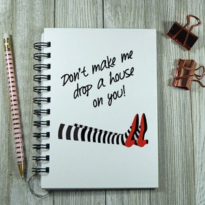 May include: A white spiral bound notebook with the text "Don't make me drop a house on you!" written in black marker. The notebook has a black and white striped leg and red shoes drawn on the front.