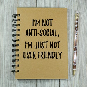 May include: A brown spiral notebook with the text "I'M NOT ANTI-SOCIAL, I'M JUST NOT USER FRIENDLY" written in black ink. A gold pencil with white snowflakes is lying on a wooden surface next to the notebook.