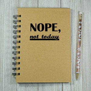May include: A brown spiral bound notebook with the text "NOPE, not today" printed on the front cover. A pencil with a white and gold design is lying next to the notebook on a light wood surface.