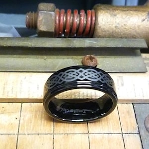 May include: A black tungsten carbide ring with a silver Celtic knot design engraved around the band. The ring is on a wooden surface.