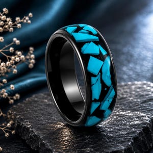 May include: A black ring featuring a wide band with an inlay of irregular, bright turquoise-colored stone fragments. The surface of the ring is smooth and polished, showcasing a high-contrast design between the dark band and the vibrant blue stone pieces.