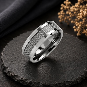 May include: A silver-toned ring featuring a central inlay with a dark gray and black checkered carbon fiber pattern. The band has polished, beveled edges that reflect light, creating a smooth, metallic appearance with a distinct woven texture.