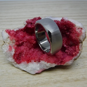 May include: A brushed silver ring with a geometric design. The ring is set against a backdrop of a white and red mineral formation. The ring's surface has a matte finish, and the inner band is polished.
