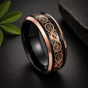 May include: A black band ring featuring a rose gold-colored inlay with an intricate, continuous Celtic-style knotwork pattern. The ring has polished black beveled edges and a smooth, dark interior, showcasing a high-contrast metallic design.