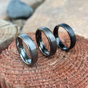 May include: Three rings with a brushed metal finish are displayed on a wooden surface. The rings vary in color, with one in silver, one in dark gray, and one in black. The rings have a simple, modern design.