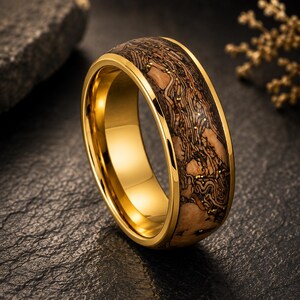 May include: A gold-toned ring featuring a polished metallic band with a wide inlay of light brown wood grain. The wood inlay is textured with dark brown, swirling organic patterns and small, scattered gold-colored flecks throughout the surface.