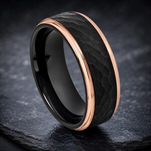 Op de afbeelding: Een ring met een brede zwarte band met een getextureerde, gehamerde afwerking. Het middengedeelte wordt geflankeerd door twee dunne, gepolijste roségoudkleurige groeven die langs de randen lopen en een contrast vormen met het donkere oppervlak.