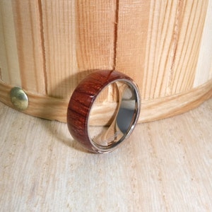 May include: A wooden and stainless steel ring with a dark red wood inlay. The ring is lying on a light wood surface.