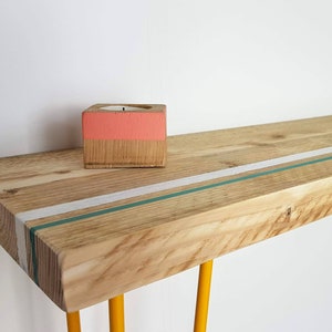 May include: A wooden shelf with a white and teal stripe painted on the top. The shelf is supported by two yellow metal hairpin legs. A small wooden candle holder with a pink top is sitting on the shelf.