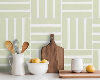 May include: A kitchen scene featuring a light green and white patterned backsplash. A white pitcher holds wooden spoons and a whisk. A wooden cutting board and a bowl of lemons are also present, along with a white bowl and a small white mug.