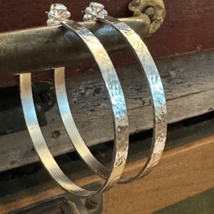May include: A pair of silver hoop earrings with a hammered texture. The earrings are thick and have a slightly rounded shape.