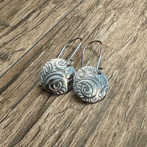 Puede incluir: Un par de pendientes de aro de plata con un diseño circular que presenta un patrón de espiral en relieve.