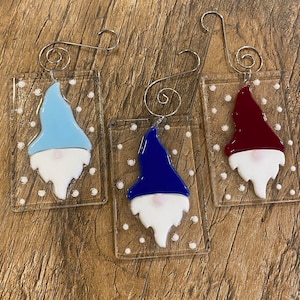May include: Three glass ornaments shaped like gnomes with white beards and different colored hats. The gnome on the left has a light blue hat, the middle gnome has a dark blue hat, and the gnome on the right has a burgundy hat. Each ornament has a silver wire hanger.
