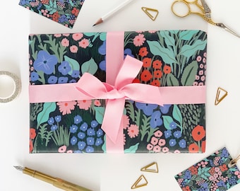 Può includere: Un regalo avvolto in carta da regalo con motivo floreale bianco e nero con un nastro rosa. Il regalo è circondato da graffette dorate, una matita, un rotolo di nastro adesivo e un paio di forbici dorate.
