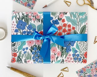 Può includere: Una carta da regalo bianca con un nastro blu e un motivo floreale con fiori rosa, rossi e blu. Il motivo include foglie verdi e forme astratte.