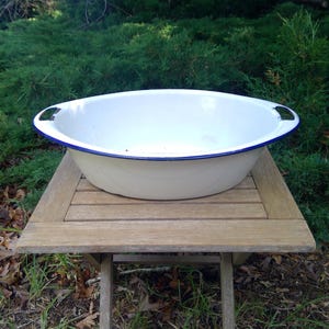 May include: An oval-shaped white enamel basin featuring a dark blue rim and two integrated side handles. The basin has a smooth, glossy interior and exterior finish, with a deep, wide bowl design suitable for holding various items or liquids.