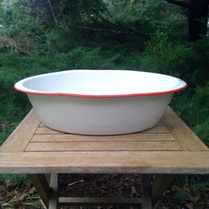 May include: A white oval enamel basin featuring a smooth finish and a thin, vibrant red rim along the top edge. The basin has a deep, rounded interior and a wide, curved exterior wall, presenting a clean and functional design for various uses.