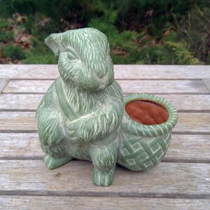 May include: A ceramic figurine of a rabbit in a sage green finish with a textured, fur-like appearance. The rabbit is standing upright next to a small, round basket with a woven pattern, featuring a contrasting terracotta-colored interior.