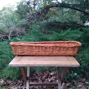 May include: A long, rectangular woven wicker basket with a natural light brown finish. The basket features a sturdy, interlaced willow construction with a flat base and slightly flared sides, designed for holding items in a horizontal arrangement.