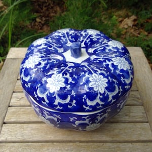 May include: A blue and white ceramic pumpkin-shaped container with a lid. The container is decorated with a floral pattern. The lid has a small stem-like handle. The container is sitting on a wooden surface.