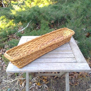 May include: A long, rectangular woven basket made of light brown wicker sits on a weathered wooden table. The basket's design features a tightly woven pattern, and it is empty. The background includes greenery and fallen leaves.