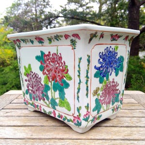 May include: A rectangular white ceramic planter featuring hand-painted floral motifs in shades of red, purple, blue, and green. The exterior displays detailed chrysanthemum blossoms and foliage, framed by thin orange lines and a decorative floral border.