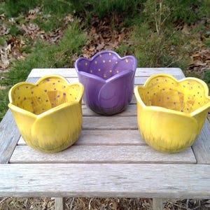 May include: Three ceramic planters shaped like tulip blossoms. Two planters are bright yellow with purple polka dots on the interior and a green brushstroke base. The center planter is purple with yellow polka dots on the interior.