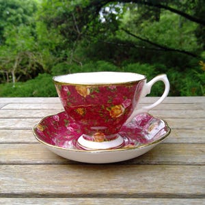 May include: A vintage teacup and saucer set. The cup and saucer are a deep red with a floral pattern in yellow and pink. The rims are trimmed with gold. The set is placed on a wooden table, with a green background.