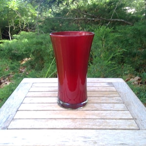 May include: A tall, red glass vase with a flared top sits on a wooden table. The vase has a glossy finish and a clear base. The background features green foliage and natural light.