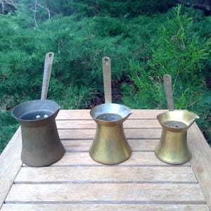 May include: Three metallic coffee pots of varying sizes arranged in a row. Each pot features a wide base, a tapered body, a pouring spout, and a long, flat vertical handle attached to the side. The pots exhibit different shades of brass and bronze.