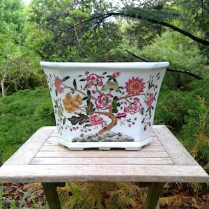 May include: A white ceramic planter with a floral design featuring pink, yellow, and red flowers. The planter is octagonal and sits on a wooden table. The background is a lush green garden.