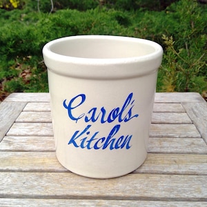 May include: A cylindrical off-white ceramic crock featuring a smooth rim and a flat base. The front of the vessel displays the text Carol's Kitchen printed in a bold, blue, cursive script font, centered vertically on the exterior surface.