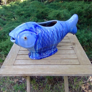 May include: A ceramic fish-shaped pitcher featuring a glossy blue glaze with darker blue accents highlighting the scale texture. The vessel has a wide open mouth at the top, a defined eye, and a sturdy base, showcasing a vibrant cobalt color palette.