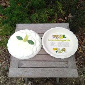 May include: A two-piece ceramic pie dish set with a scalloped edge. The lid features a sculpted lemon wedge and two green leaves. The base interior displays a printed recipe for Lemon Meringue Pie with illustrations of lemons, eggs, and butter.