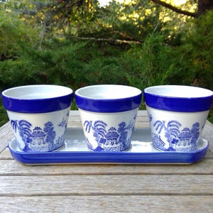 May include: Three white ceramic planters with a blue willow pattern and a solid blue rim, arranged in a row on a matching rectangular white tray with a blue border. Each planter features a detailed blue landscape illustration of trees and buildings.