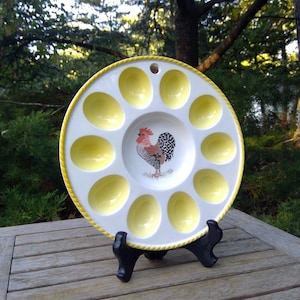 May include: A white ceramic egg serving platter with a yellow border and egg-shaped indentations. The center features a rooster illustration. The platter is displayed on a black stand, set against a natural outdoor backdrop.