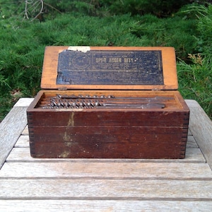 May include: A rectangular wooden box with a hinged lid containing a set of metal auger bits. The interior of the lid features a black label with the text "SPUR AUGER BITS" printed in white. The box has visible finger joints at the corners.