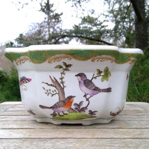 May include: A white ceramic planter with a scalloped rim featuring a green and gold textured border. The exterior is decorated with hand-painted illustrations of two birds perched on branches, a small green mound, and colorful butterflies.