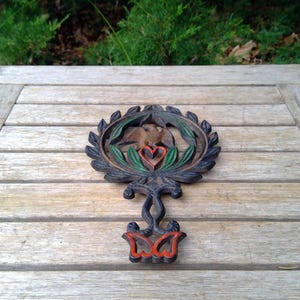 May include: A cast iron trivet with a decorative design featuring a bird, heart, and leaf motif. The trivet is painted in shades of brown, green, and red. The trivet is resting on a wooden table.