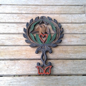 May include: A dark-toned cast iron trivet featuring a circular frame with a laurel leaf border. Inside the circle, a central brown bird figure sits above a red heart. The base of the trivet has a decorative handle with red accents at the bottom.