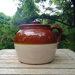 May include: A round ceramic crock with a glossy brown glazed upper section and a matte cream-colored lower section. The vessel features a single side handle and a matching brown lid with a small knob handle centered on the top surface.