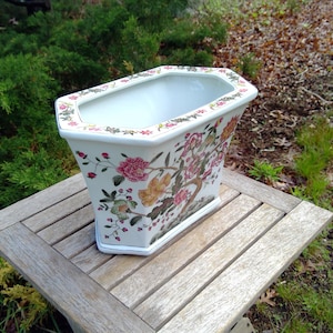 May include: A white, octagonal ceramic planter with a floral design in pink, orange, and green. The planter sits on a wooden table outdoors, surrounded by greenery and foliage. The rim of the planter is decorated with a floral border.