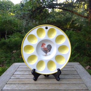 May include: A white ceramic egg serving platter with a yellow rim and egg-shaped indentations. The center features a rooster illustration. The platter is displayed on a black stand against a natural outdoor backdrop.