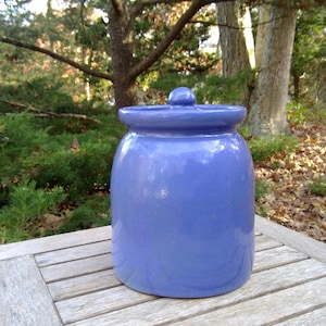 May include: A cylindrical ceramic storage jar with a smooth, glossy periwinkle blue glaze. The jar features a wide, flat rim and a matching circular lid topped with a small, rounded knob handle, presenting a uniform and solid color finish.