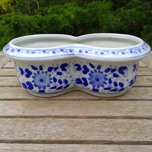 May include: A white ceramic planter with a distinctive figure-eight shape. The planter is adorned with blue floral patterns and a blue trim along the rim. The planter is sitting on a wooden table.