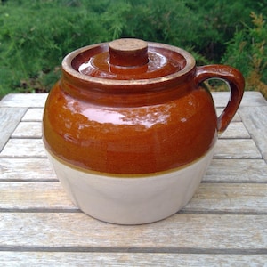 May include: A two-toned ceramic crock featuring a glossy brown glazed upper section and a matte cream-colored base. The vessel includes a matching brown lid with a central knob handle and a single side handle for gripping the round container.
