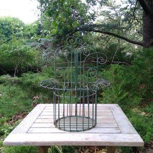 May include: A green metal plant stand with a decorative, scrolled design. The stand has a circular base and a cage-like structure, perfect for supporting climbing plants. It is placed on a wooden table outdoors, surrounded by lush greenery.