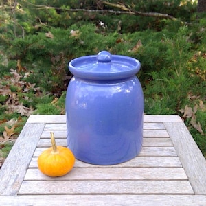May include: A smooth, periwinkle blue ceramic canister with a matching lid featuring a small knob handle, positioned next to a small, round, orange pumpkin on a slatted wooden surface.